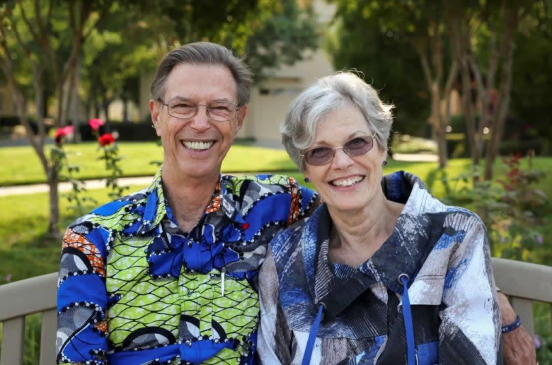 An older man and woman sit closely together on a bench outdoors, smiling. Both wear colorful patterned shirts and glasses. Green trees and flowers are visible in the background.