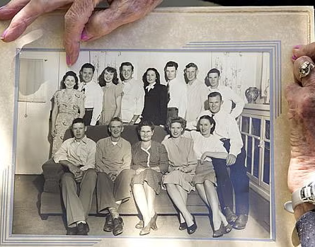 Two elderly hands hold a vintage black-and-white photo showing a large family, with five people seated in front and eight people standing behind them, all posing indoors.