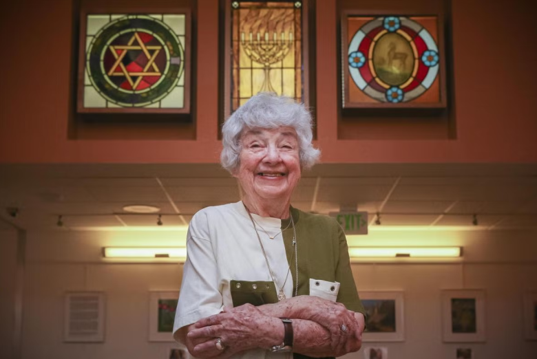 An elderly woman with gray hair smiles and stands with arms crossed in front of stained glass panels featuring religious symbols in a warmly lit room.