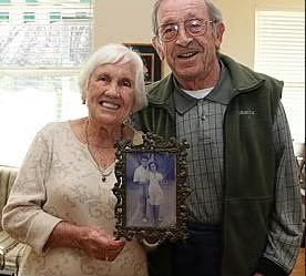 An elderly smiling couple stands together indoors while holding a framed old photograph of a younger couple, possibly themselves in their youth.