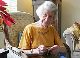 An elderly woman with white hair sits in an armchair, knitting. She wears a yellow shirt and looks toward the camera. The room is cozy, with a plant, a cushioned chair, and soft lighting in the background.