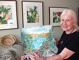 An elderly woman with long white hair smiles while holding a colorful landscape painting. Behind her, four framed floral paintings hang on the wall. A woven basket sits nearby.
