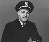 A man in a dark naval officers uniform and cap poses for a formal portrait against a plain background.