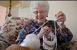 An elderly woman with white hair and glasses sits on a floral armchair, smiling as she knits with white yarn. A lamp is visible in the background.
