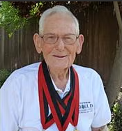 Elderly man with glasses smiles outdoors, wearing a white shirt and three gold medals with red and black ribbons around his neck.