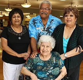 Four older adults, three standing and one seated in a wheelchair, smile warmly in a well-lit indoor setting. The group appears to be enjoying each others company.