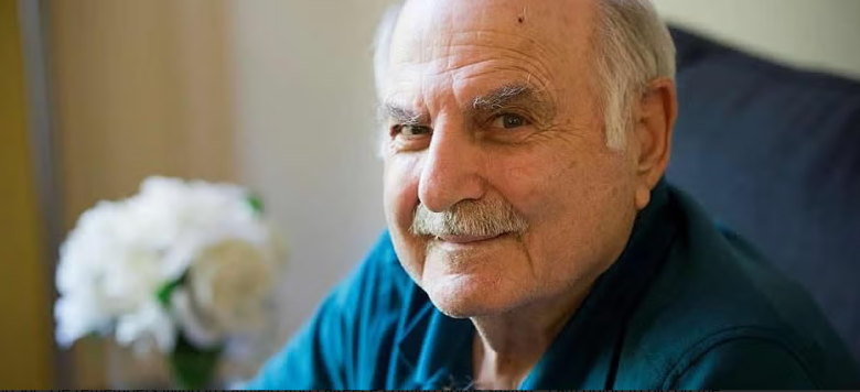 An elderly man with white hair and a mustache smiles while sitting indoors. He is wearing a blue shirt, and white flowers in a vase are visible in the blurred background.