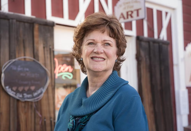 A smiling woman with short, curly brown hair wears a teal sweater and stands in front of a rustic building with a neon open sign and visible wood doors.
