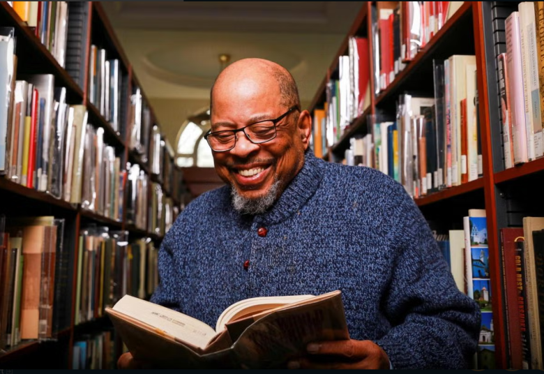 A smiling man wearing glasses and a blue sweater stands between library bookshelves, holding and reading an open book. Light shines through a window behind him.