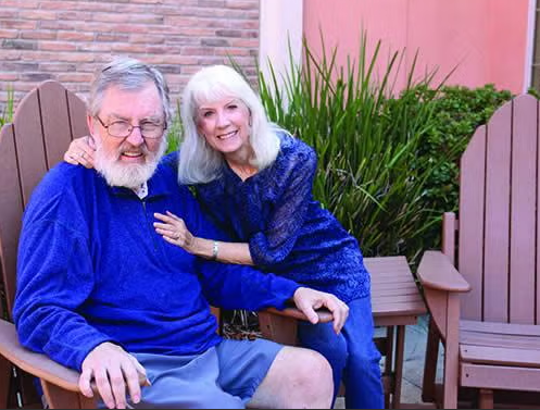 An older man and woman sit together on wooden chairs outside. The man wears a blue sweater and glasses, while the woman, also in blue, smiles with her arm around his shoulder. Green plants and a brick wall are in the background.