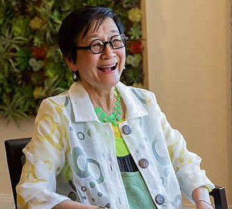 A smiling woman with short black hair and glasses wears a white, patterned jacket and green necklace, sitting indoors with green plants in the background.