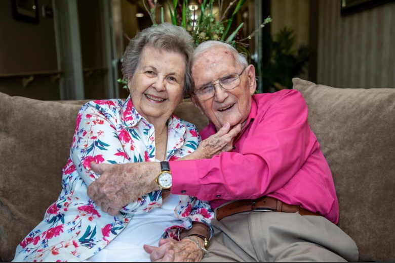 An elderly couple sits closely together on a couch, smiling warmly. The woman wears a floral blouse and the man wears a bright pink shirt. They appear happy and content, with her arm gently around his shoulders.
