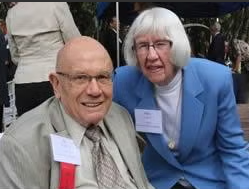 An elderly man in a suit and an elderly woman in a blue blazer are smiling outdoors at what appears to be a formal event. Both wear name tags. Other people are visible in the background.