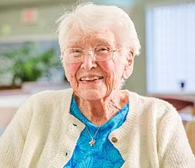 Smiling elderly woman with white hair, glasses, and a cream-colored sweater over a blue top, sitting indoors with a blurred background.