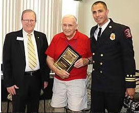 Three men pose indoors; one, wearing red and holding a plaque, stands between a man in a suit and a man in a firefighter dress uniform.