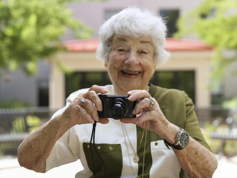 Smiling elderly woman with white hair holds a camera outdoors, wearing a two-tone shirt and jewelry, as if ready to capture the next chapter in her tai chi tale, with greenery and a building blurred in the background.