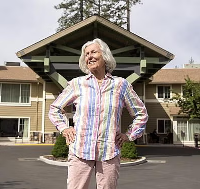 An older woman with gray hair smiles confidently outside a modern building, wearing a colorful striped shirt and light pants, as if savoring the feeling of coming home. Trees and a clear sky create a welcoming backdrop.