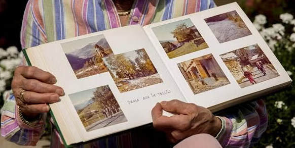 A person wearing a colorful striped shirt holds an open photo album displaying several vintage outdoor photos, each evoking a warm sense of coming home. The background includes blurred greenery and white flowers.