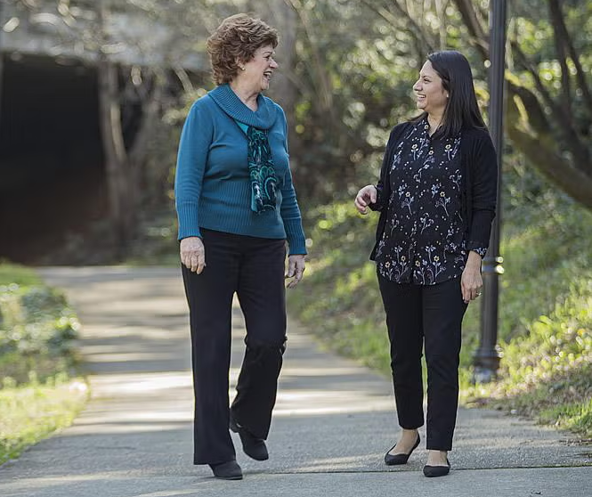 Two women walk and talk on a paved path in a park, surrounded by trees and greenery, dressed in casual clothing and appearing to enjoy their conversation as if catching up after coming home.