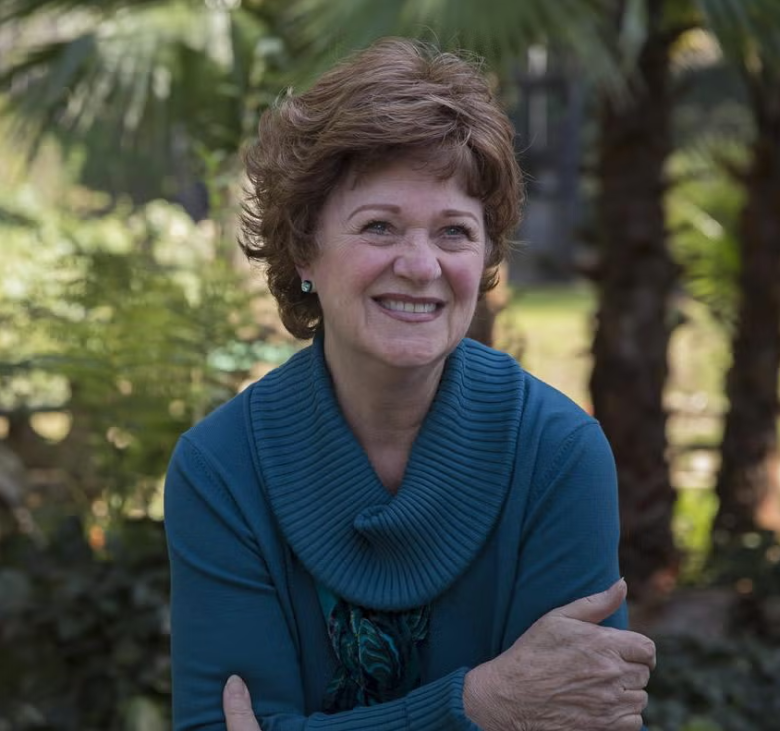 A woman with short curly hair, wearing a teal sweater, smiles while standing outdoors in front of palm trees and greenery, capturing the warm, joyful essence of coming home.