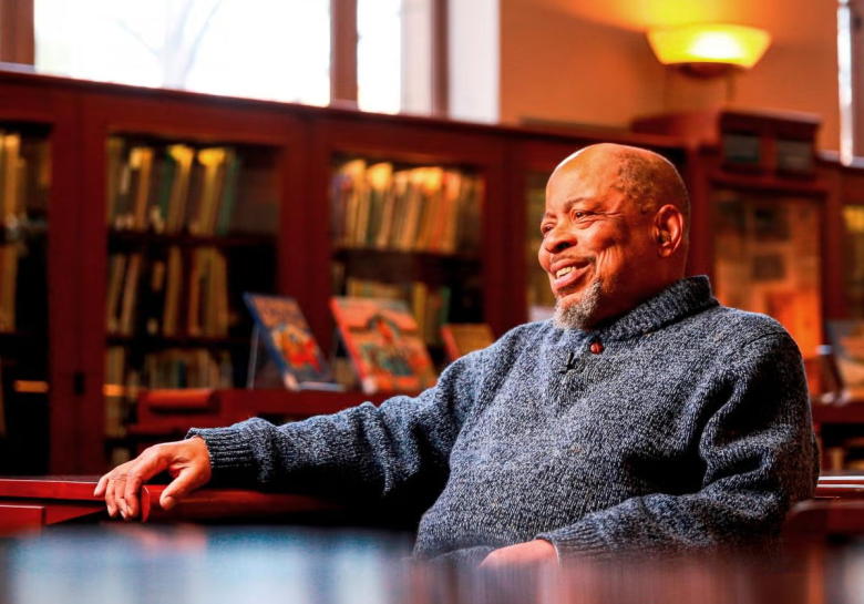 A smiling man with a beard sits in a library wearing a blue sweater, surrounded by bookshelves and soft lighting in the background, as if pausing thoughtfully on his own Journey of the Heart.