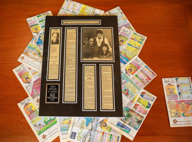 A framed newspaper article titled “Journey of the Heart,” with photos and text, is placed on a wooden table, surrounded by colorful comic or puzzle pages arranged in a circular pattern around the frame.