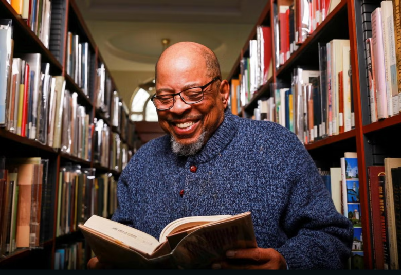 A smiling man in glasses and a blue sweater reads a book titled Journey of the Heart while standing between bookshelves in a library.