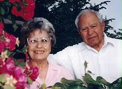 Meet Lucy: An elderly woman and man stand close together outdoors, smiling, surrounded by greenery and pink flowers. The man wears a white shirt; the woman—meet Lucy—wears glasses and a light pink blouse.