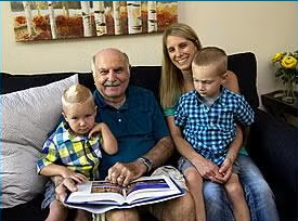 An elderly man sits on a couch with two young boys and a woman, all smiling as they enjoy their time together. The man holds an open book on his lap, ready to share a story. Meet Lucy, who brings warmth to the scene with her cheerful presence. A painting and some flowers are visible in the background.