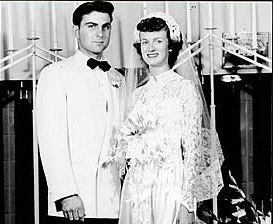 A bride in a long-sleeved lace wedding dress and veil stands smiling next to a groom in a white tuxedo jacket and black bow tie. Meet Vic, who appears to be posing with his bride for a formal wedding portrait.