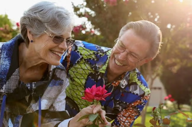 Two older adults wearing glasses and colorful shirts smile together in a sunlit garden, admiring a blooming red rose. Their joyful expressions reveal a close bond, and it’s clear that this pair loves to dance through life’s beautiful moments.