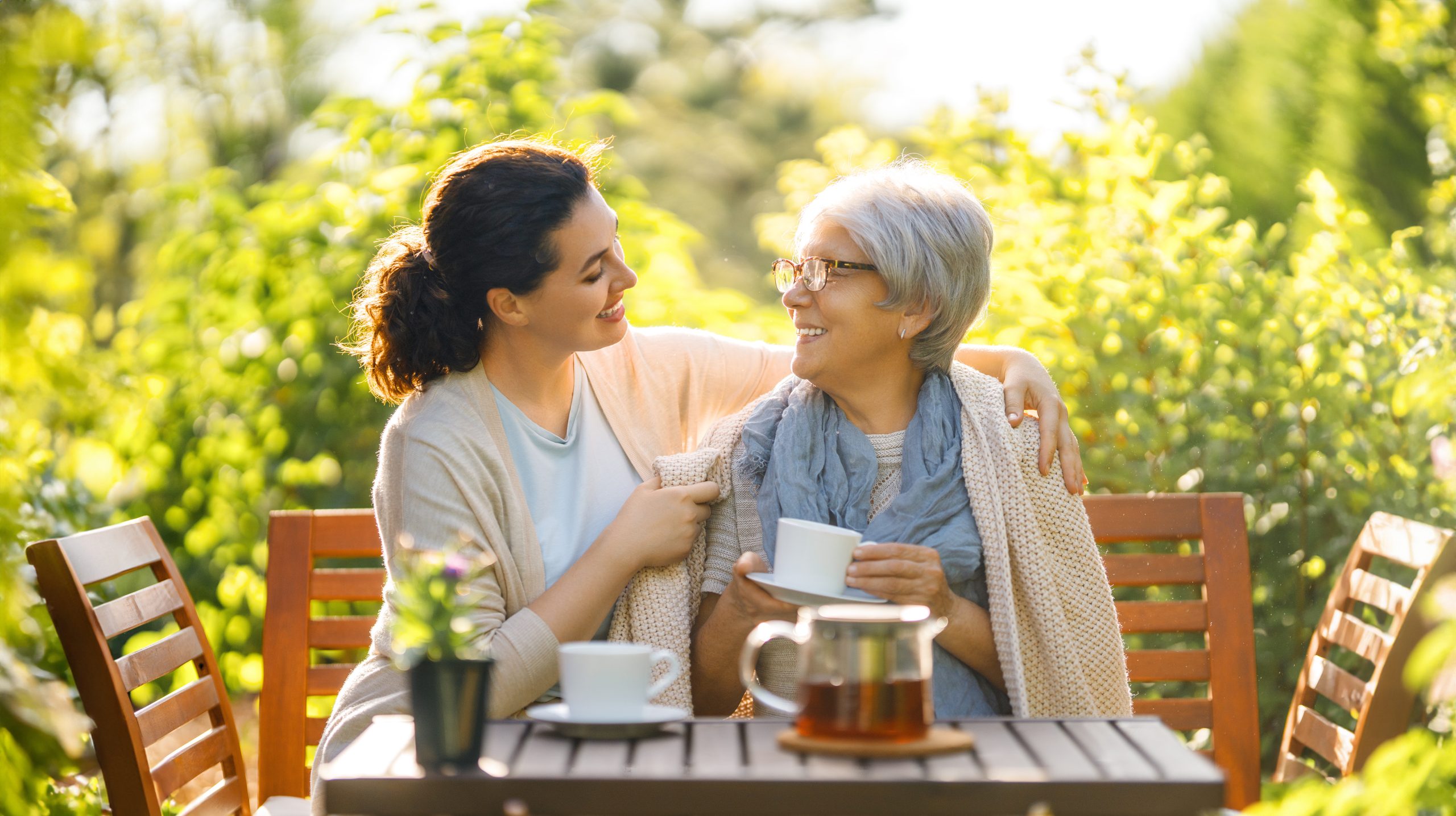 A young woman smiling at an older woman as she helps her wrap a blanket around her shoulders while they sit at an outdoor table with tea.